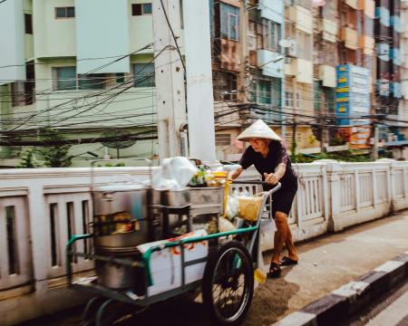 Thailändischer Mann beim Schieben seines Verkaufswagens mit Essen auf einem Gehsteig.
